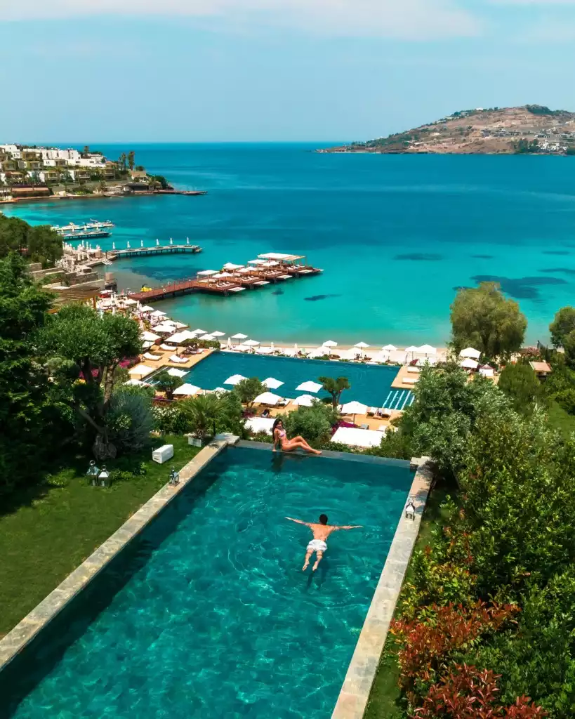Bodrum'un turkuaz sularına nazır lüks bir tesisin sonsuzluk havuzunda sırtüstü yüzen bir erkek ve kenarında uzanan bir kadınla, şemsiyeli plaj alanı ve ahşap iskelelerin yer aldığı berrak koyun kuşbakışı, cenneti andıran bir manzarası.
