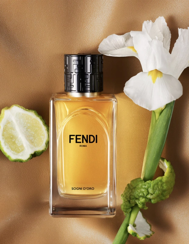 Şeffaf cam şişedeki altın rengi Fendi Sogni D'Oro parfümü, üzerinde markanın logosu desenli siyah kapağıyla sıcak kahverengi zeminde sergileniyor; yanında yarım bergamot meyvesi ve yeşil sapına bergamot kabuğu sarılmış beyaz bir iris çiçeği bulunuyor.