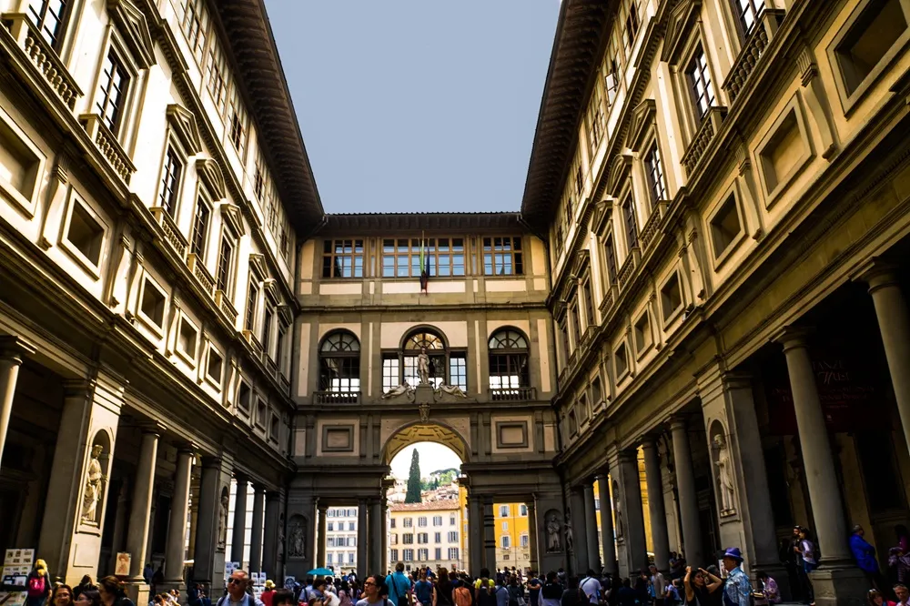 Floransa, İtalya'daki Uffizi Galerisi'nin geniş U-şekilli avlusunda, klasik mimariye sahip görkemli binaların kolon ve heykellerle süslü cepheleri arasında gezen kalabalıklar ve kemerli geçidin ötesinde uzanan şehir manzarası belirgin.