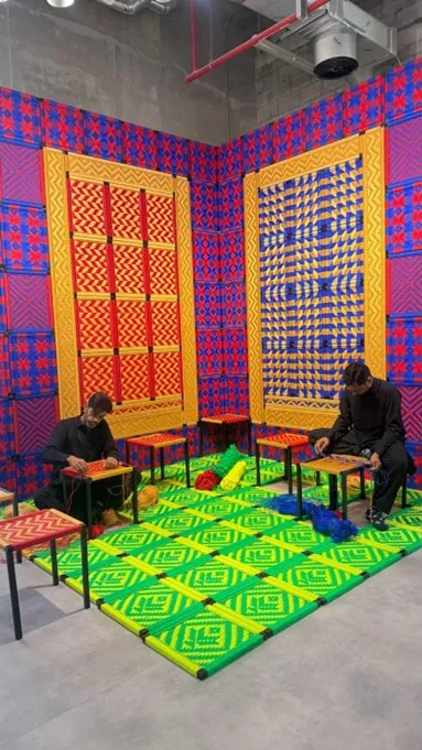 Art Basel Katar’da Imran Qureshi’nin Nature Morte başlıklı çalışmasında, canlı renklerde dokuma yüzeyler ve masa başında üretim yapan katılımcılar yer alıyor.
