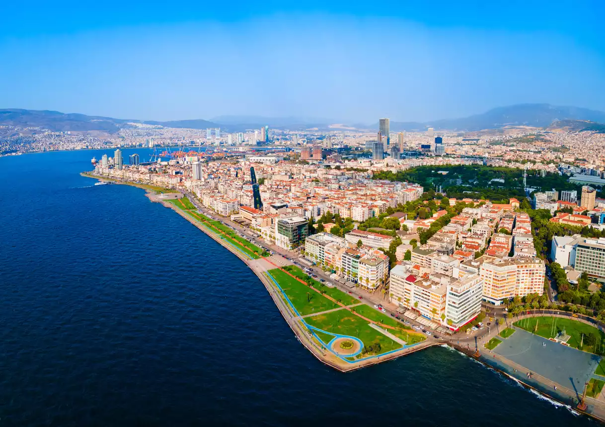 Berrak masmavi bir gökyüzünün altında, İzmir Körfezi'nin derin mavi sularının yemyeşil sahil şeridi parklarıyla buluştuğu, çağdaş mimariye sahip yüksek binaların ve yoğun şehir dokusunun kuşbakışı panoramik görüntüsü.