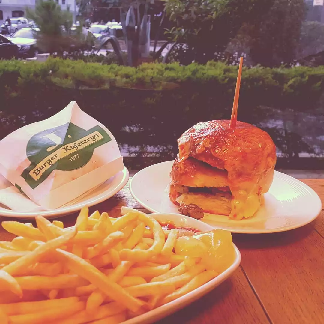 Duru Akgül'ün şeflerin lezzet rotaları kapsamında favori hamburger mekanlarından J Burger'da çekildiği anlaşılan bu yakın planda, erimiş peynirleri akan büyük bir hamburgerin yanında bol porsiyon patates kızartması ve üzerinde Burger Kafeterya 1977 yazan bir paket ahşap masada iştah açıcı duruyor.