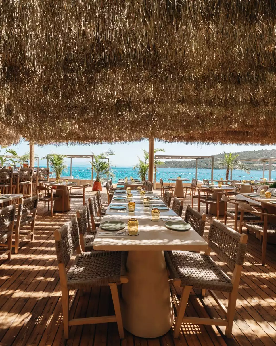 Kuum Hotel'in denize sıfır konumunda bulunan, doğal hasır çatılı ve ahşap zeminli, şık örgülü sandalyelerle donatılmış açık hava restoranında, masmavi Ege Denizi manzarasına karşı özenle hazırlanmış masalar eşliğinde huzurlu bir yemek alanı.