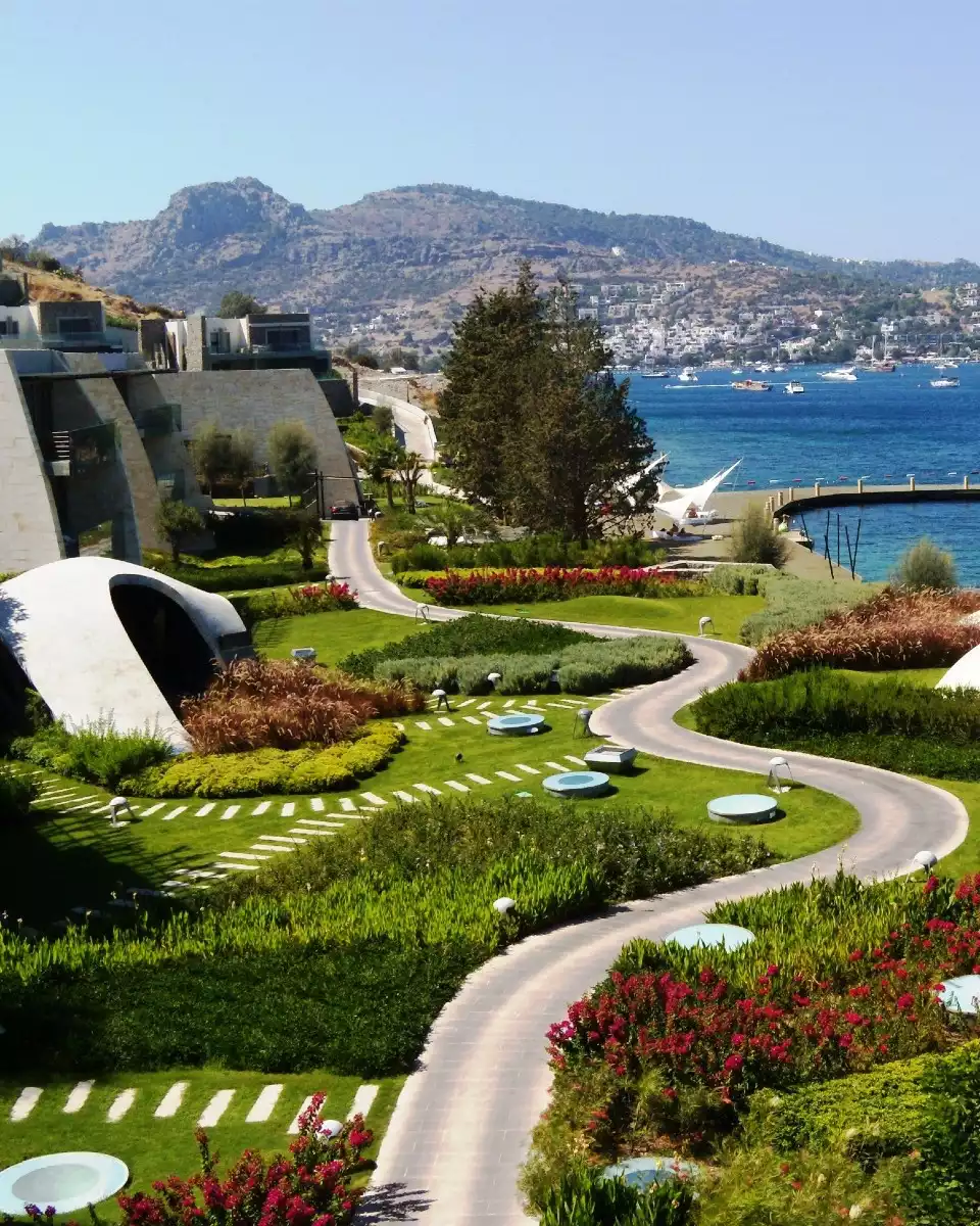 Kuum HotelSpa'nın yemyeşil peyzajlı ve modern mimarili yapılarını çevreleyen, kıvrımlı yolları ve rengarenk çiçeklerle bezeli bahçelerinin önünden görünen; masmavi denizin, teknelerin ve karşıdaki dağlık alana yayılan yerleşim yerinin huzurlu birleştiği bir sahil panoraması.