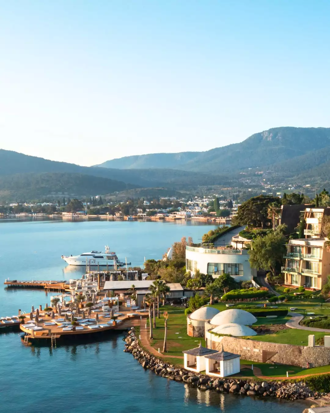 Kuum Hotel & Spa'nın Bodrum'daki nefes kesen konumu, mavi denize uzanan iskelesi, şezlonglarla dolu dinlenme alanları, modern mimarisi, yemyeşil bahçeleri ve uzak dağlarla çevrili sakin atmosferini kuş bakışı gösteren bir görüntü.