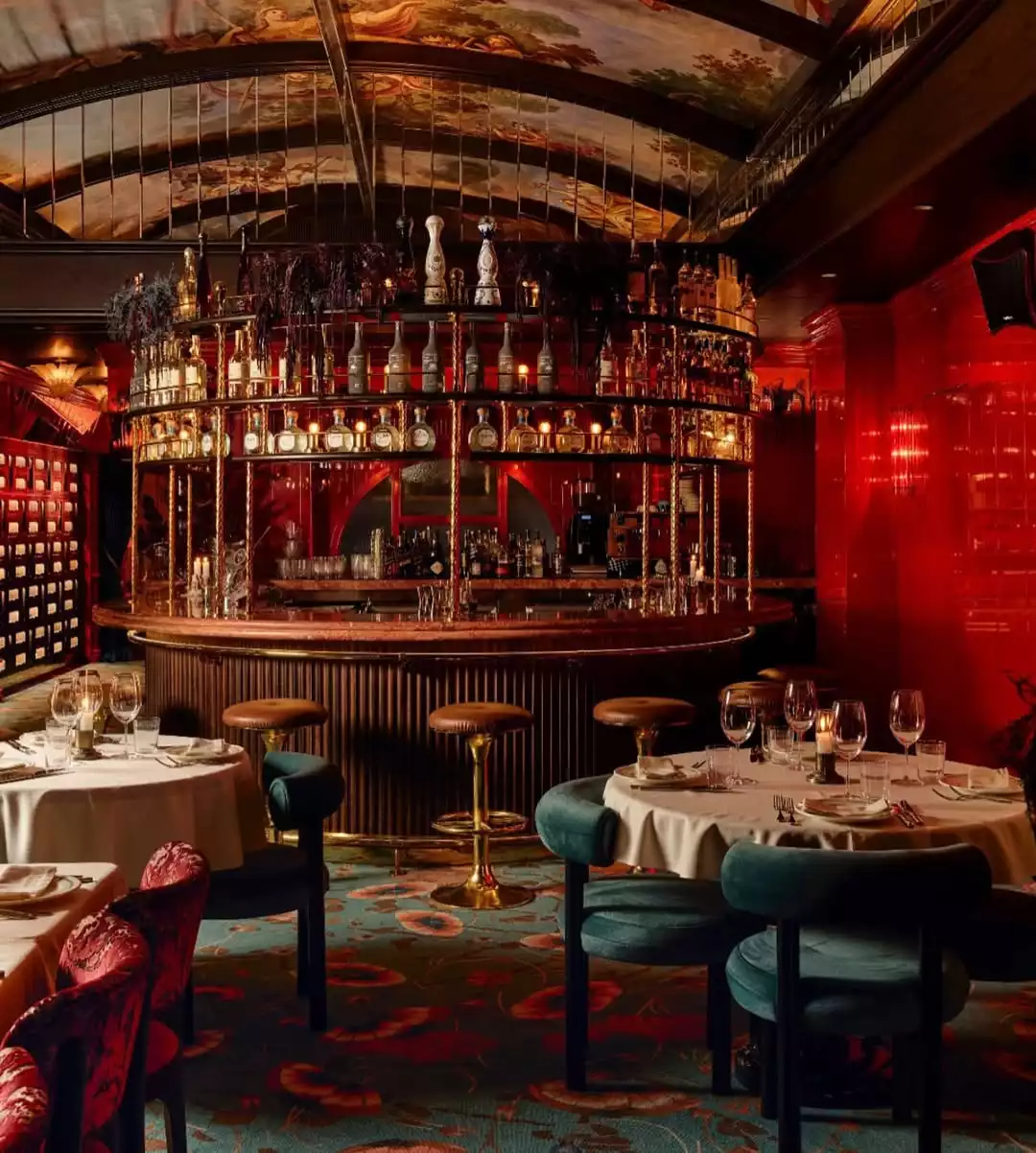 Loş ışıklarla aydınlatılmış, kırmızı duvarlara ve karmaşık desenli tavana sahip lüks bir bar veya restoran iç mekanında; arka planda çok sayıda şişenin sergilendiği ahşap ve metal detaylı görkemli bir bar, ön planda ise kadife görünümlü koyu renkli sandalyelerle çevrili, beyaz örtülü şık yuvarlak masalar bulunmaktadır.