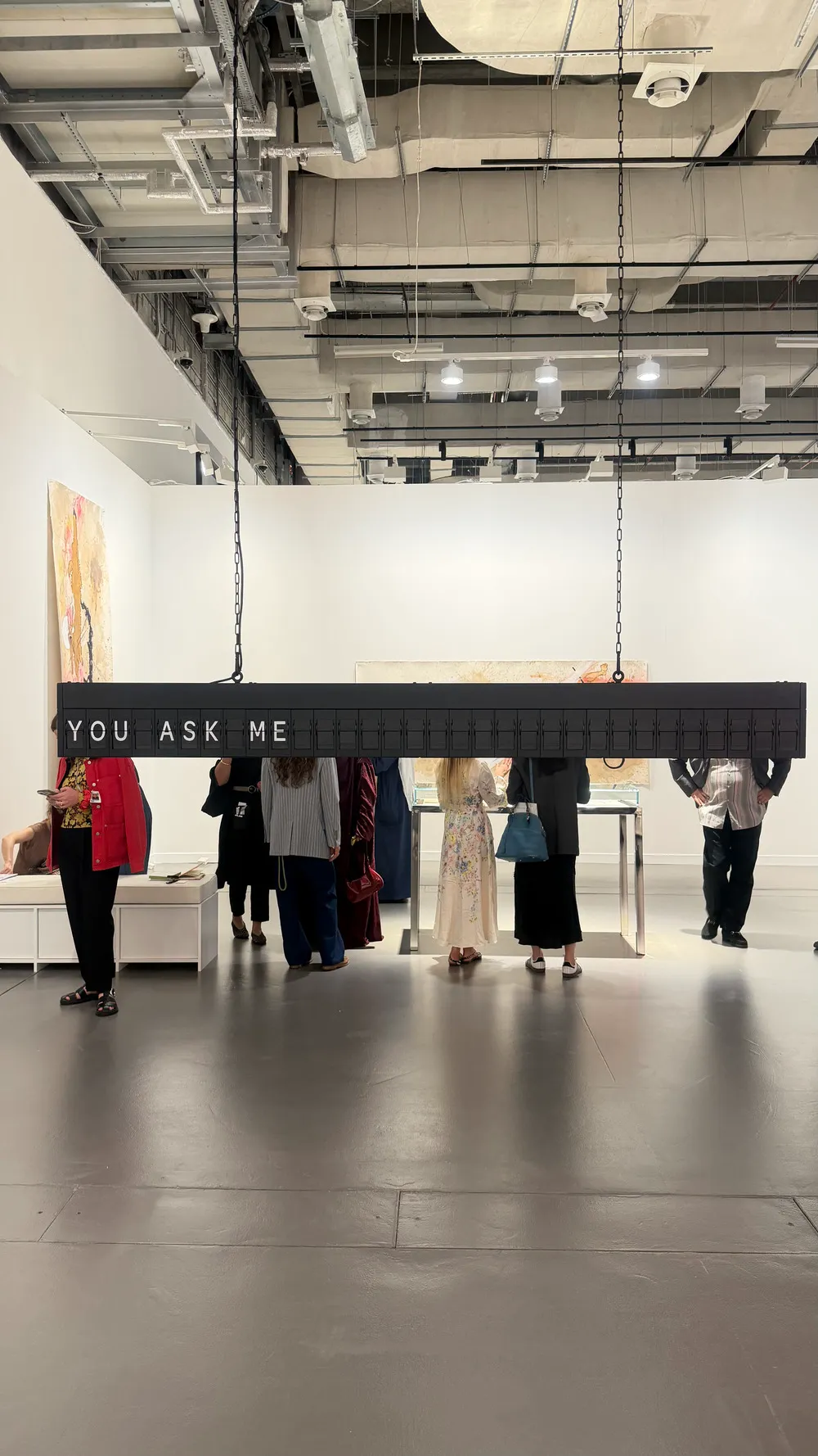 Art Basel Katar’da Shilpa Gupta’ya ait, tavandan zincirlerle asılı ve “YOU ASK ME” ifadesini oluşturan metin yerleştirmesi önünde ziyaretçiler yer alıyor.