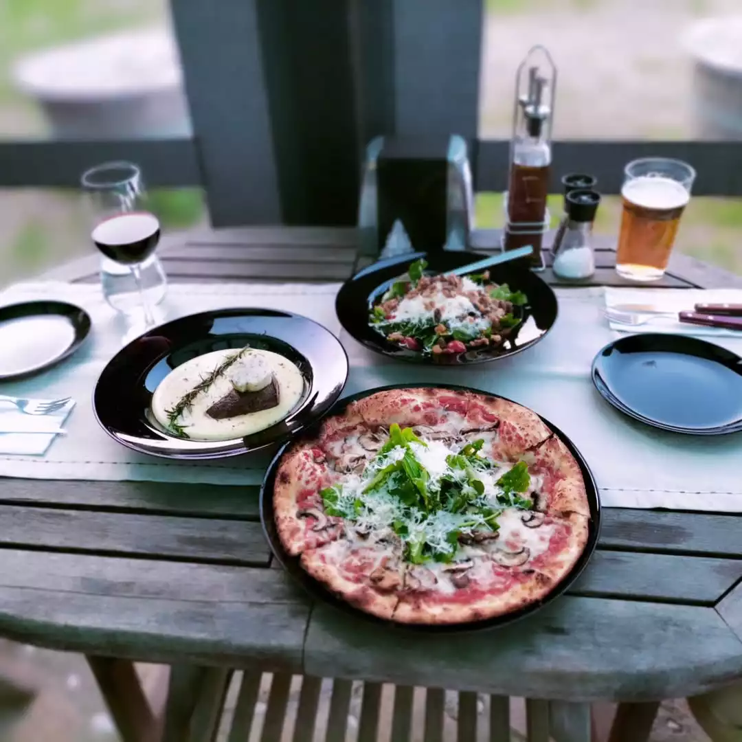 Datça'daki Trio Pizza gibi şef Duru Akgül'ün Şeflerin Lezzet Rotaları başlığı altında keşfetmeye değer bulduğu bir duraktan, mantarlı, roka ve bol rendelenmiş peynirli iştah açıcı pizzanın, kremalı soslu et yemeğinin ve yeşillik salatasının sunulduğu, kırmızı şarap ve bira ile tamamlanmış şık bir açık hava masası.