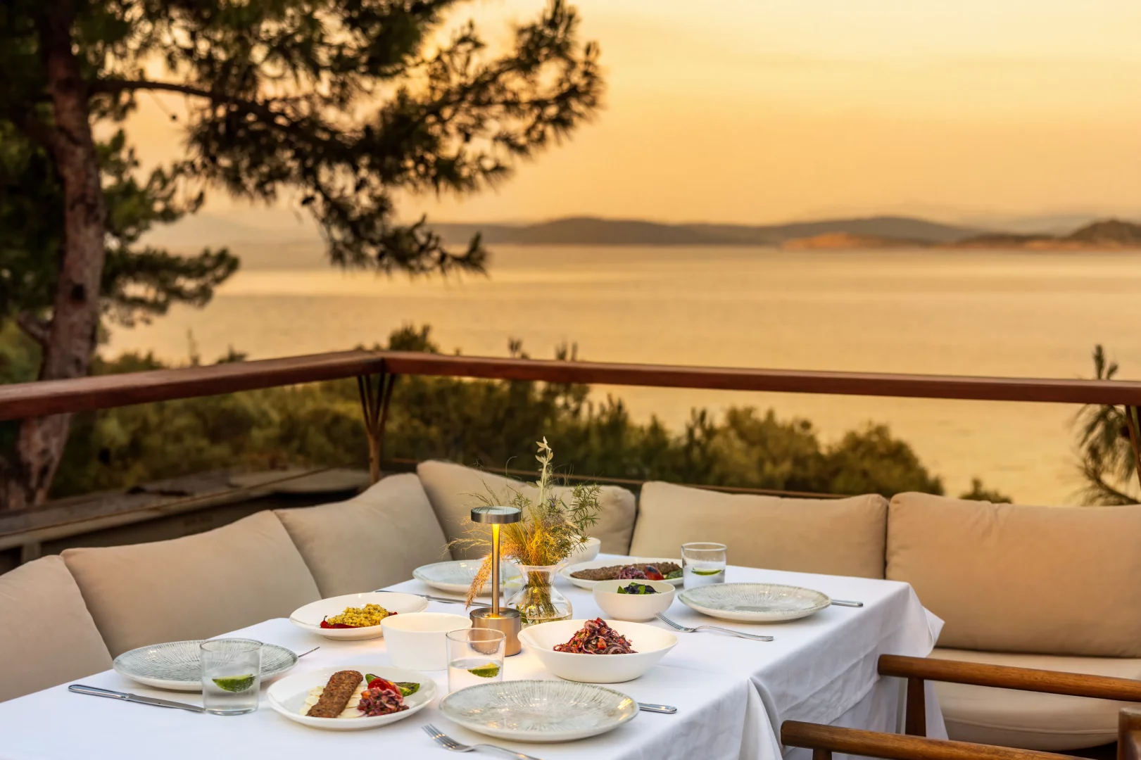 Gün batımının denize yansıdığı, ağaçlarla çevrili bir terasta, beyaz masa örtülü ve bej minderli koltuklarla düzenlenmiş, üzerinde kebap, rengarenk mezeler ve içeceklerin bulunduğu şık bir akşam yemeği masası.