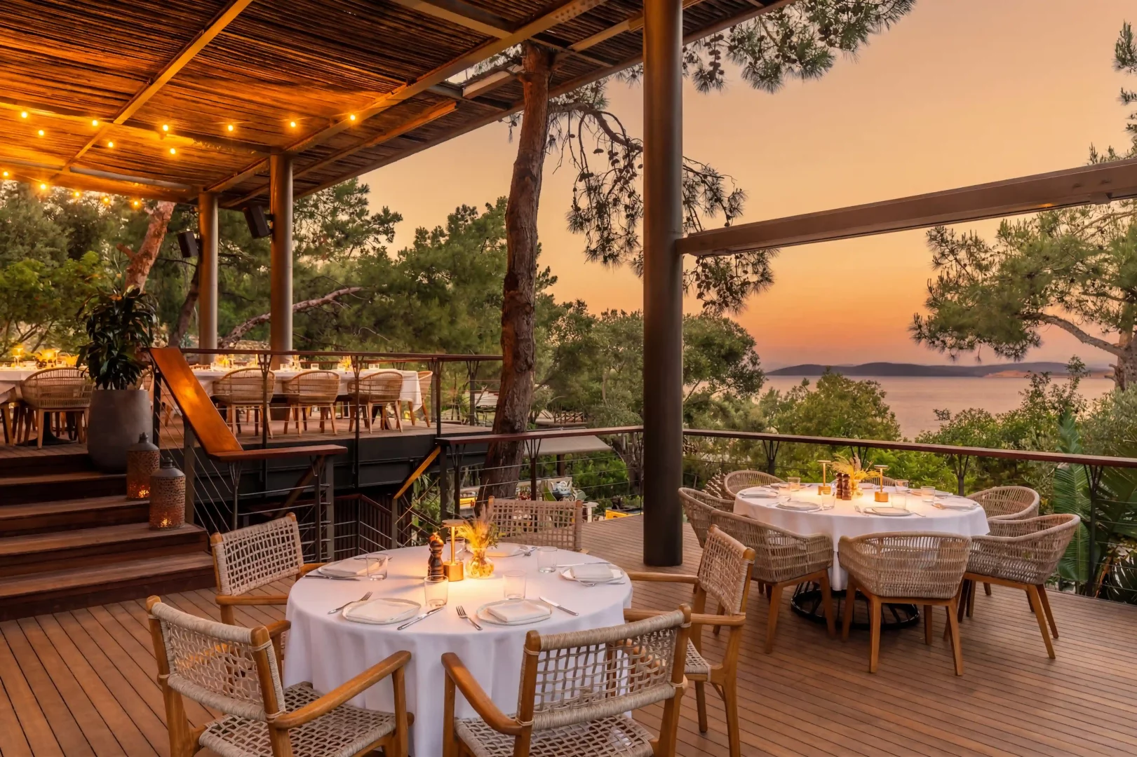 Gün batımının turuncu ve pembe tonlarıyla aydınlanan gökyüzünün altında, masaları özenle hazırlanmış, ahşap teraslı şık bir açık hava restoranı, uzakta görünen deniz manzarası ve çevresindeki yeşilliklerle huzurlu bir yemek deneyimi sunuyor.