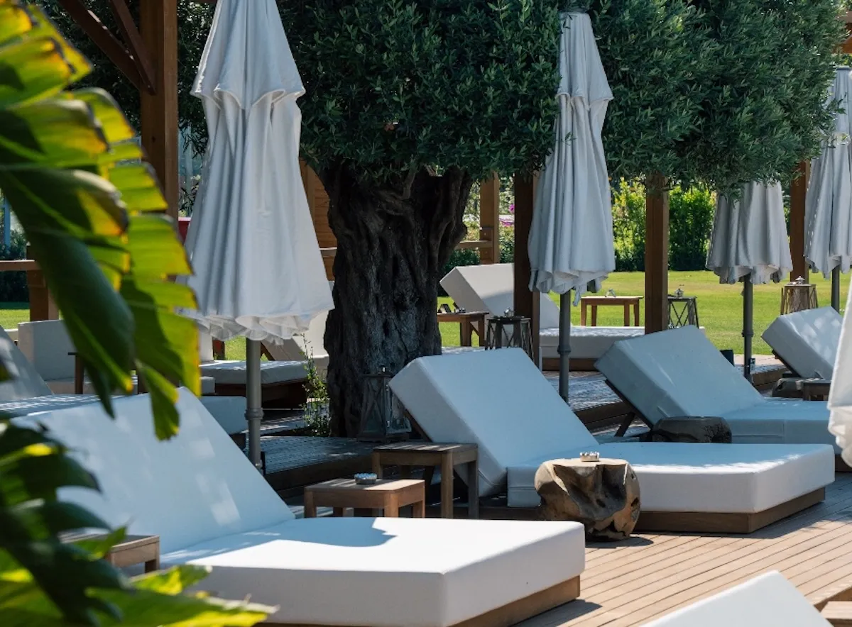 Akra Antalya Oteli'nin yemyeşil bahçesinde, ahşap zemine sıralanmış beyaz minderli şezlonglar ve kapalı şemsiyeler, çevresindeki zeytin ağaçlarının gölgesinde huzurlu bir dinlenme alanı sunuyor.