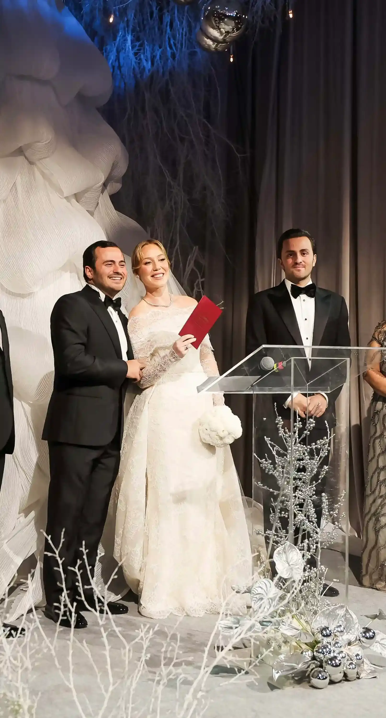 Nikâh töreninde kırmızı evlilik cüzdanını tutan gelin ve damat; dantel gelinlik, siyah smokin ve zarif sahne dekorasyonu eşliğinde resmi düğün anı.