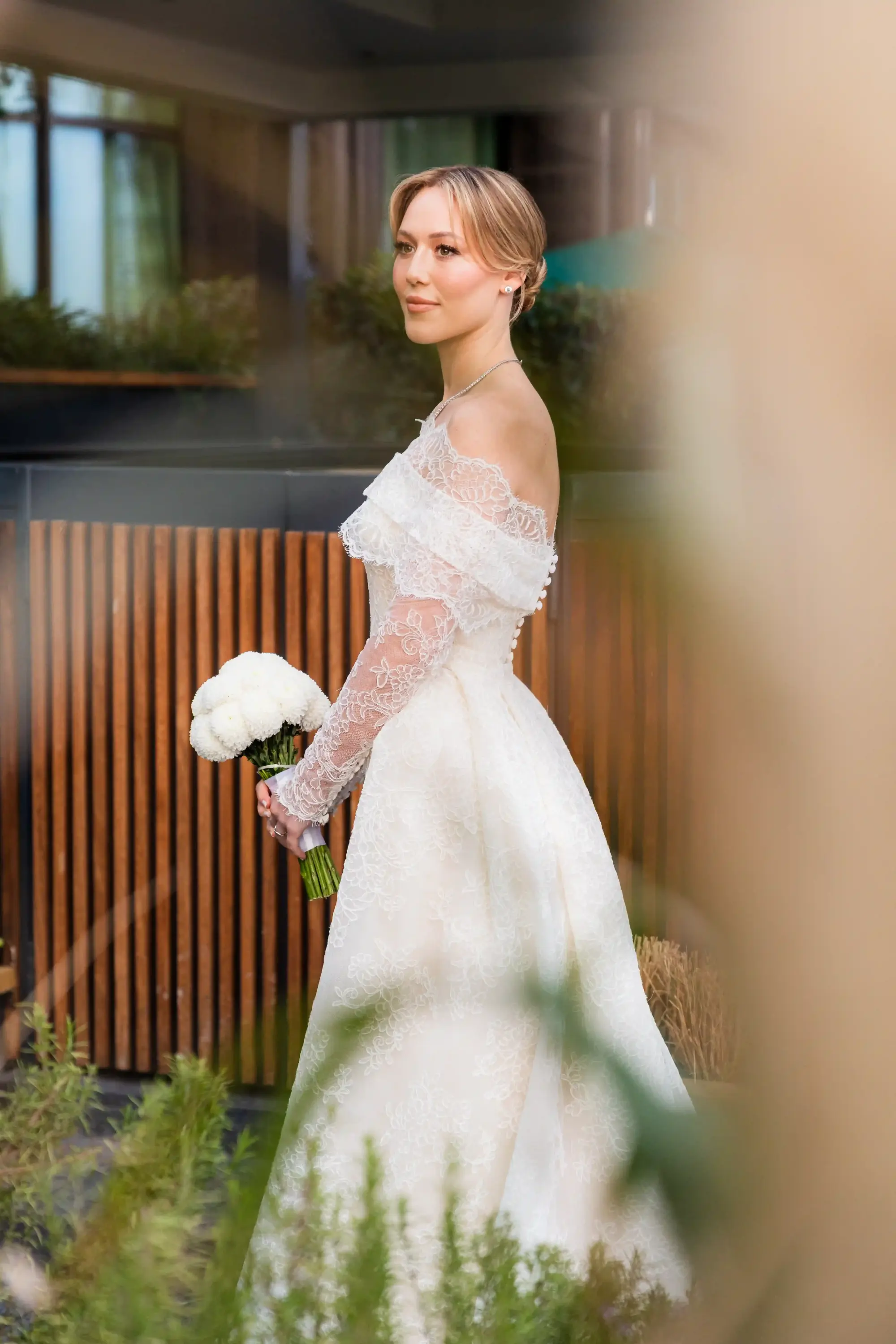 Dantel detaylı off-shoulder gelinlik giyen gelin, doğal gelin makyajı ve beyaz çiçek buketiyle açık havada yapılan zarif düğün çekiminde romantik bridal stilini yansıtıyor.