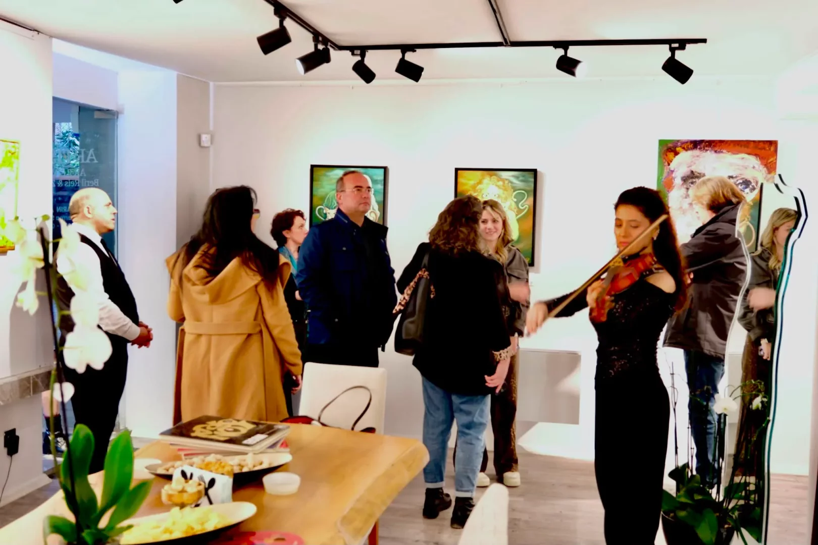 Sanat galerisi açılışında, siyah elbiseli bir kadının keman çaldığı, çeşitli soyut tabloların raylı spot ışıkları altında sergilendiği, ziyaretçilerin etrafta dolaştığı ve ön planda bir ikram masasının bulunduğu canlı bir atmosfer.
