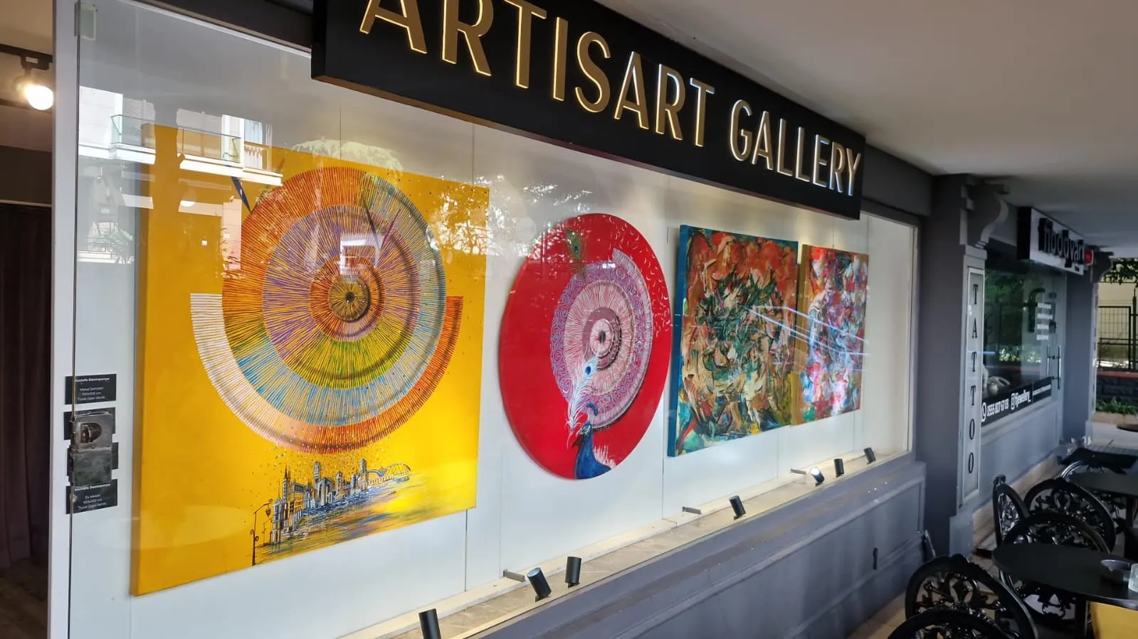 Artisart Galeri'nin vitrininde, sarı zemin üzerinde gökkuşağı renklerinde spiral desenli ve şehir silüeti içeren büyük bir tablo, kırmızı dairesel zemin üzerinde tavus kuşu motifli bir eser ve soyut formların hakim olduğu iki renkli sanat eseri, alttan aydınlatmalarla vurgulanarak sergileniyor.
