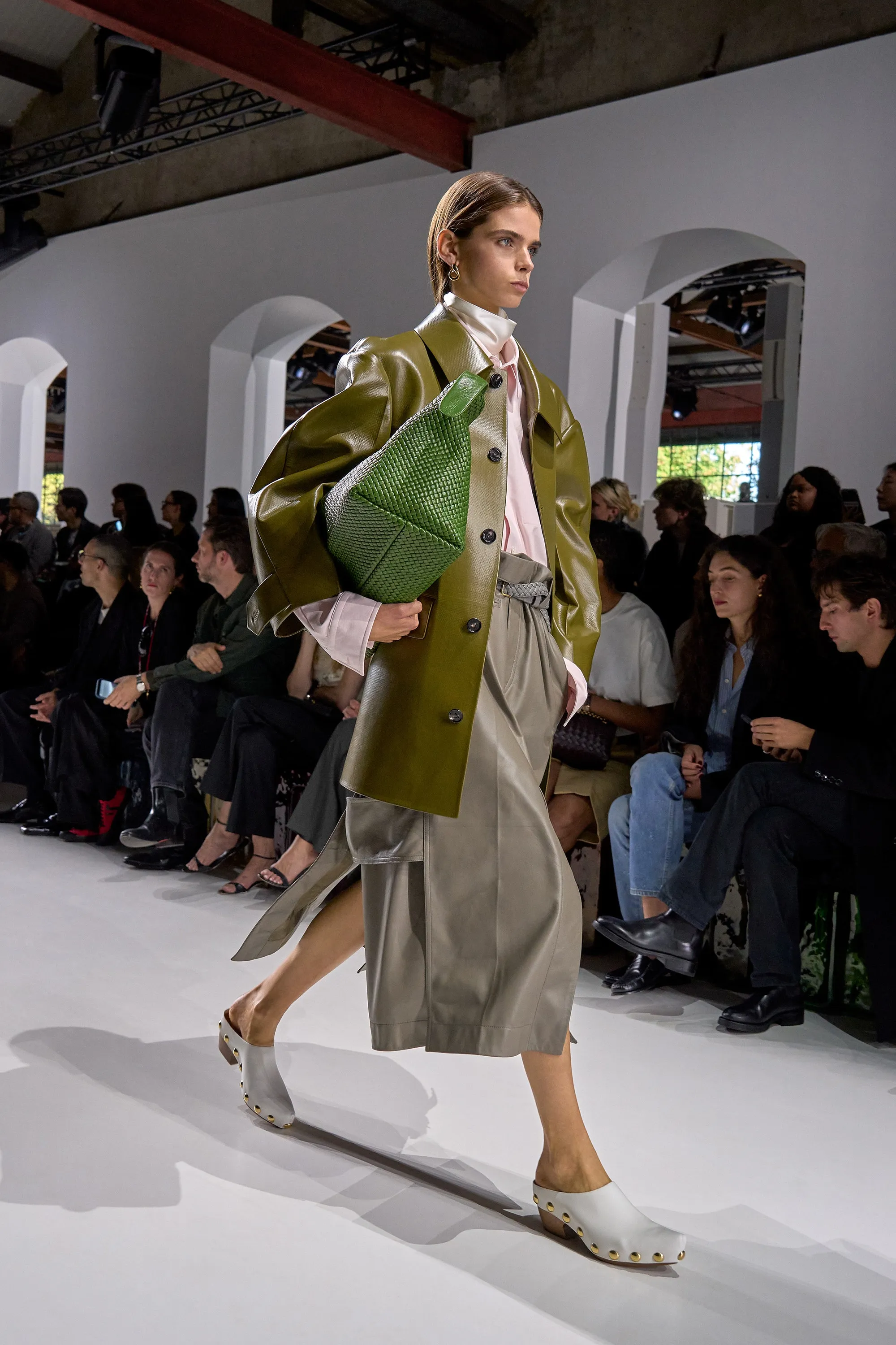 Bottega Veneta İlkbahar-Yaz 2026 defilesinde model, zeytin yeşili deri ceket ve akışkan midi etek kombinini örgü clutch çanta ile tamamlayarak modern tailoring ve güçlü, sofistike bir siluet sergiliyor.