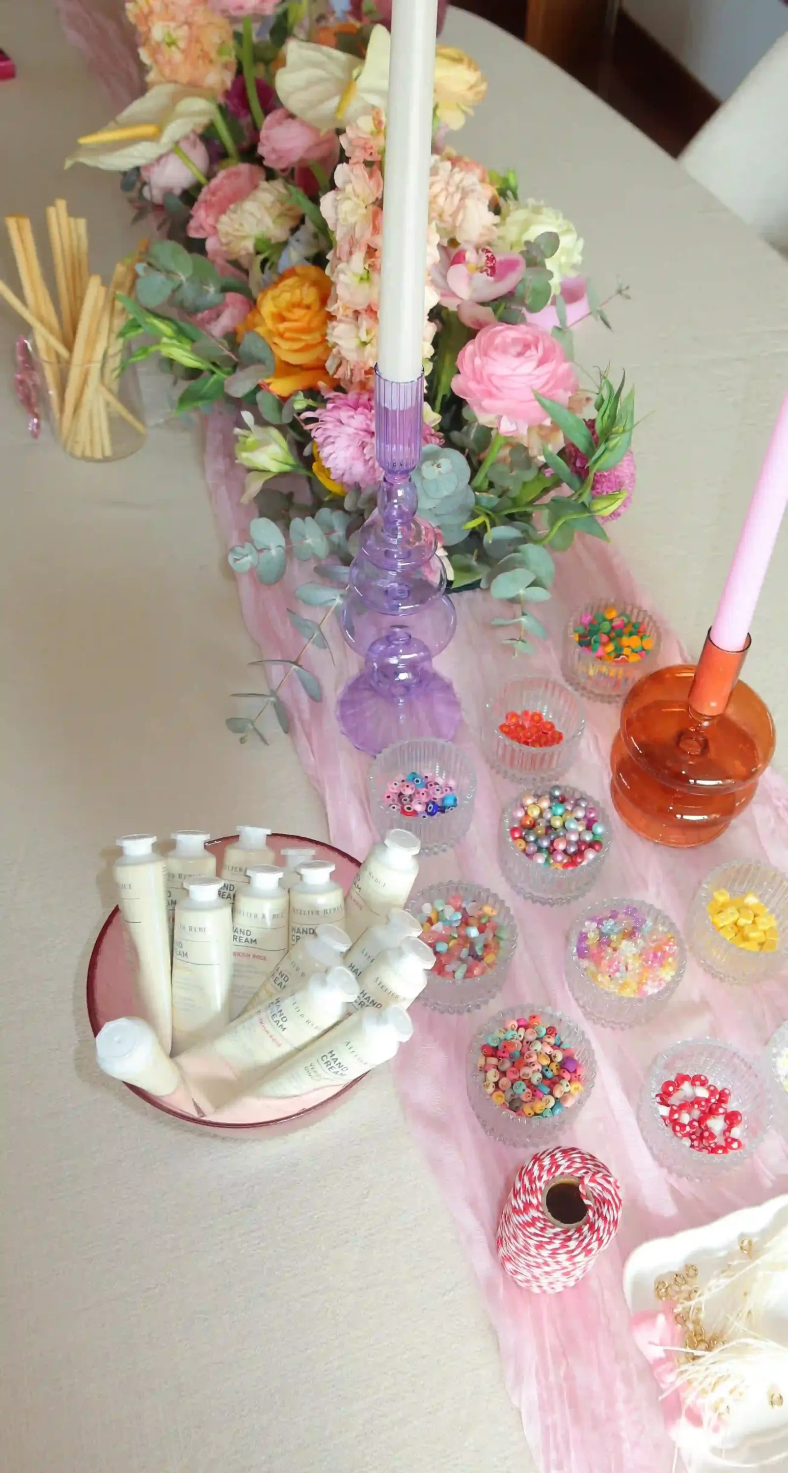Bridal shower etkinliğinde hazırlanan renkli boncuklar, charm aksesuarları, el kremi hediyeleri ve pastel tonlarda çiçek aranjmanlarıyla dekore edilmiş zarif ve yaratıcı masa düzeni.