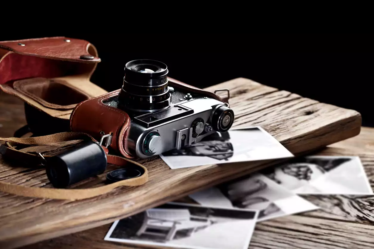 Rustik ahşap bir zemin üzerinde duran, eski tip bir film fotoğraf makinesi, yanında kahverengi deri kılıfı ve etrafa dağınık bir şekilde saçılmış, nostaljik görünümlü siyah beyaz basılı fotoğraflarla, fotoğrafçılığın geçmişini yansıtan bir atmosfer sunuyor.