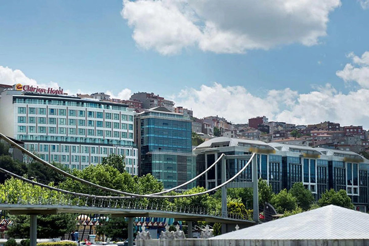 Modern mimarisiyle dikkat çeken Clarion Hotel binası, Haliç bölgesinin yeşil ağaçlarla çevrili kentsel dokusu içinde, ön planda estetik bir köprü yapısıyla ve arka planda tarihi dokulu yamaç evleriyle gökyüzünün altında uzanıyor.