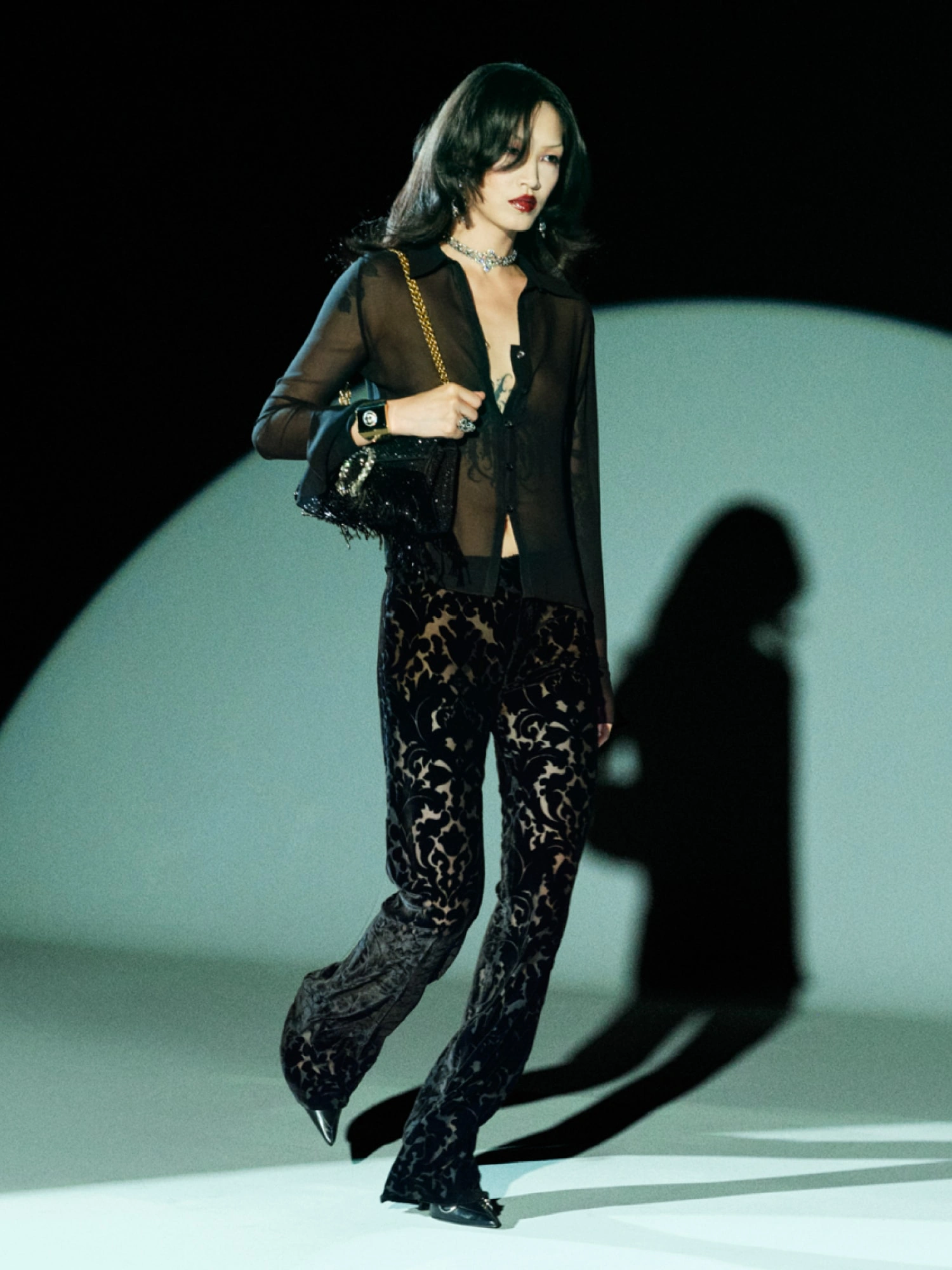 Demna’nın Gucci koleksiyonundan transparan siyah gömlek ve desenli kadife pantolon giyen model, zincir askılı çanta ve güçlü styling ile modern, cesur ve sofistike bir podyum görünümü sergiliyor.