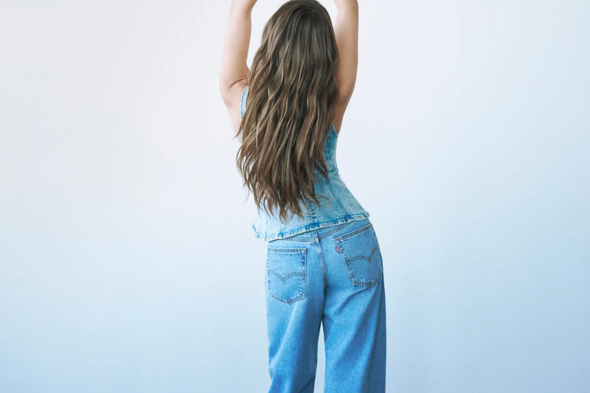 Açık mavi denim yelek ve yüksek bel jean pantolon giyen kadın modelin arkadan görünümü; uzun dalgalı saçları ve sade açık renk stüdyo fonuyla minimal moda çekimi.