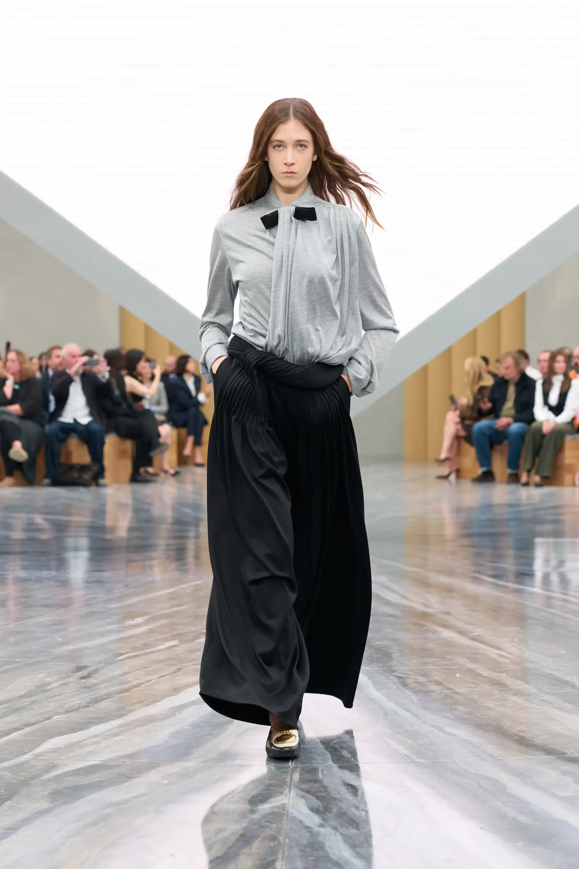 Dior İlkbahar-Yaz 2026 defilesinde model, gri drapeli bluz ve yüksek bel akışkan siyah wide leg pantolon ile modern tailoring ve zarif silueti öne çıkaran sofistike bir runway görünümü sergiliyor.