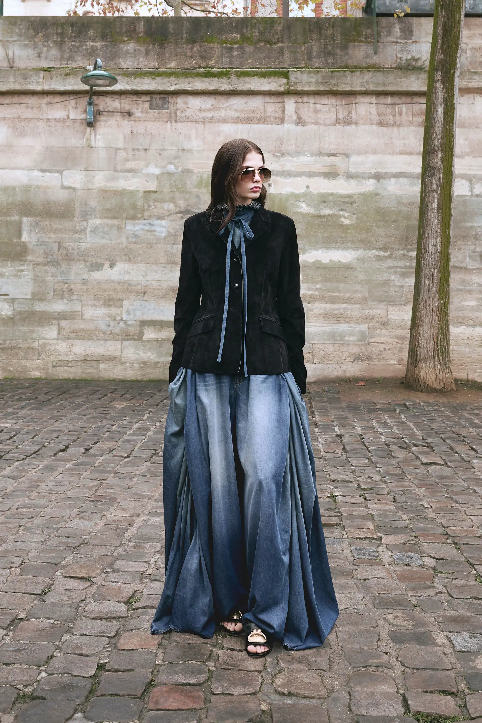 Dior Sonbahar-Kış 2026 koleksiyonundan siyah kadife blazer ve akışkan denim efektli wide leg pantolon giyen model, taş zeminli Paris sokaklarında modern tailoring ve güçlü silueti yansıtan editoryal bir görünüm sergiliyor.