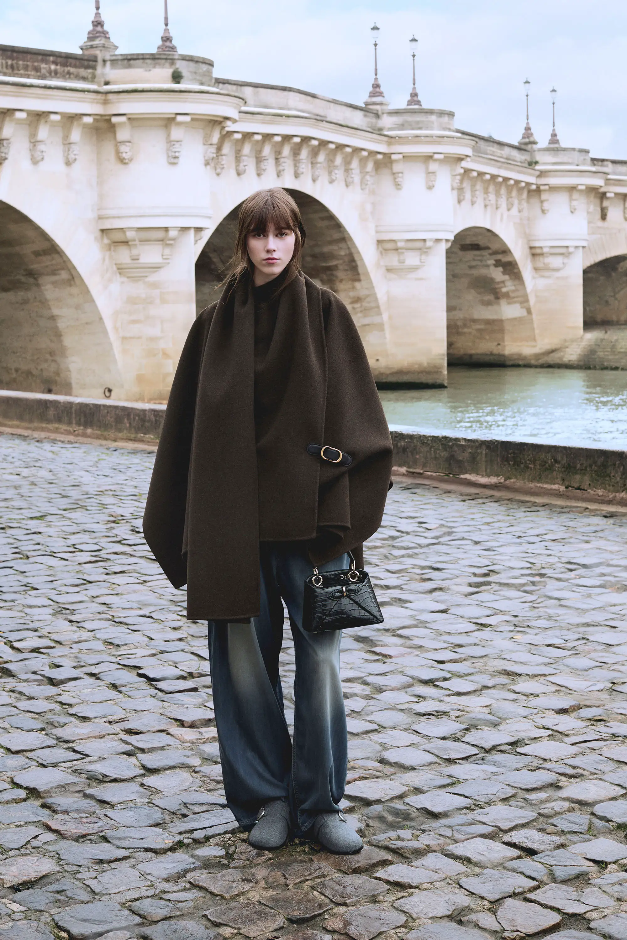 Dior Sonbahar-Kış 2026 koleksiyonundan kahverengi oversize pelerin ve akışkan wide leg pantolon giyen model, Paris’te köprü önünde zamansız, sofistike ve güçlü silueti vurgulayan modern bir görünüm sunuyor.