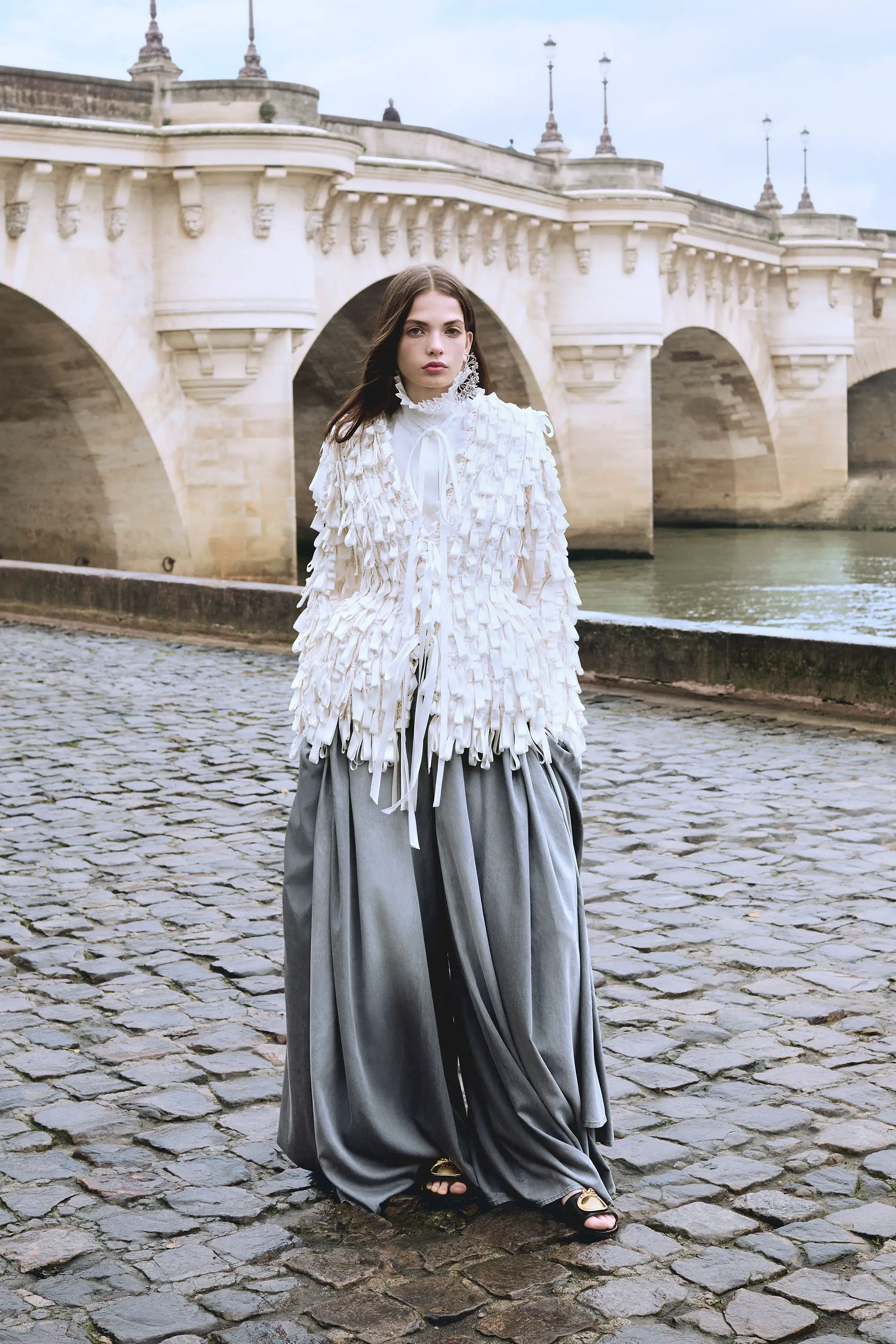Dior Sonbahar-Kış 2026 koleksiyonundan beyaz dokulu üst ve akışkan gri wide leg pantolon giyen model, Paris’te taş köprü önünde modern romantizmi ve güçlü silueti yansıtan editoryal bir görünüm sergiliyor.