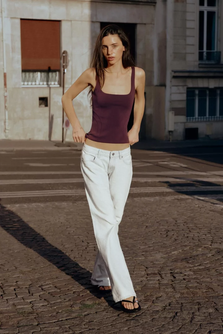 Kaldırımda duran, bordo kare yaka atlet ve açık renk düşük bel jean giymiş genç bir kadın, elleriyle pantolonunun belini hafifçe aşağı çekerek 2026 jean trendleri arasında öne çıkan düşük bel kesimin modern Y2K etkisini vurguluyor.