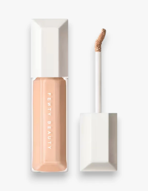Beyaz arka plan üzerinde konumlandırılmış, Fenty Beauty markasına ait açık bej tonlarda nemlendirici ve uzun süre kalıcı kapatıcının şeffaf cam şişesi, üzerinde FENTY BEAUTY yazısıyla dikkat çekiyor; hemen yanında ise ucu aynı tonda ürünle kaplı, kullanıma hazır aplikatörü ayrı olarak görülüyor.