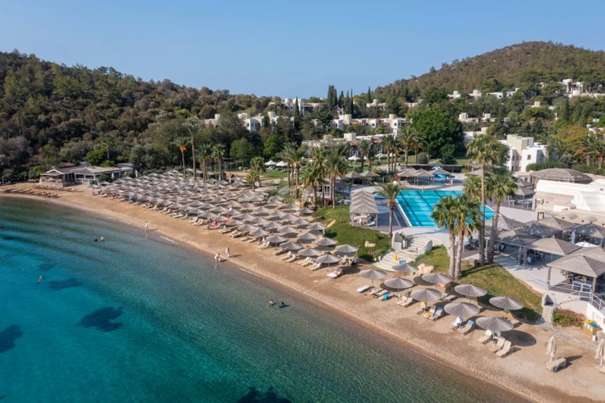 Hapimag Sea Garden Resort Bodrum'da, berrak masmavi denizin kıyısındaki geniş kumsalda gölgelik sunan birçok şemsiye ve şezlong, palmiye ağaçlarıyla çevrili ışıl ışıl bir yüzme havuzu ve yemyeşil tepelere yayılmış tesis binaları güneşli bir günde kuşbakışı gözleniyor.