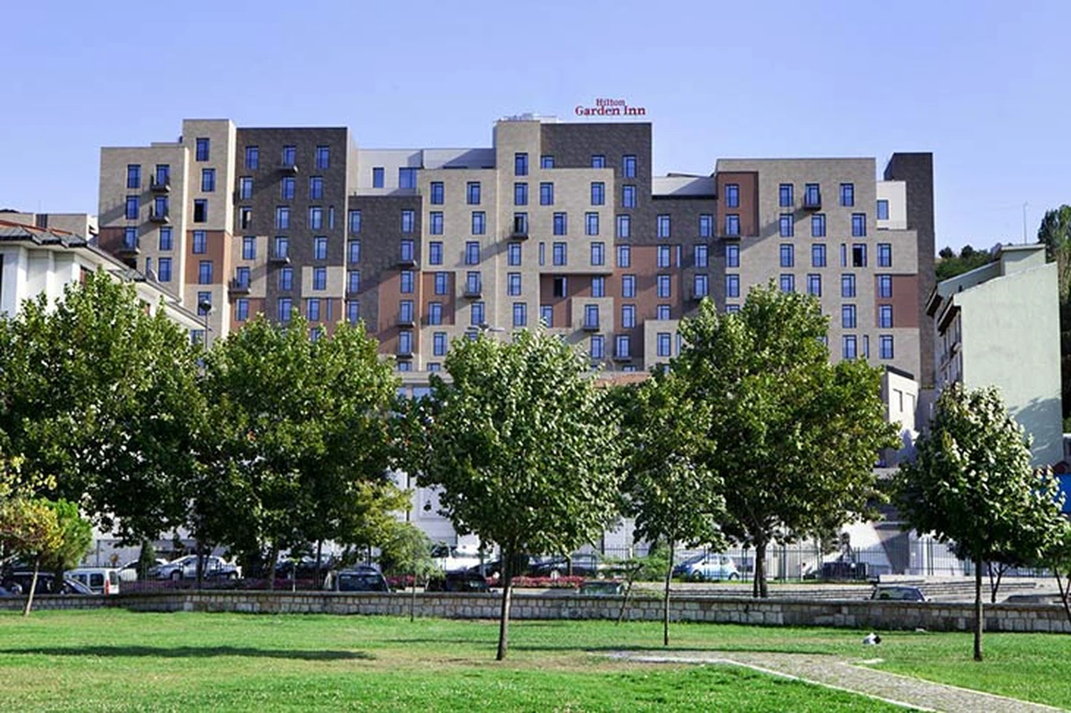Hilton Garden Inn Golden Horn otelinin modern mimarili, bej, koyu gri ve kahverengi tonlardaki çok katlı cephesi ile çok sayıda pencere ve balkonu ön plana çıkarken, ön kısımda yemyeşil bir park ve ağaçlar masmavi gökyüzünün altında yer alıyor.