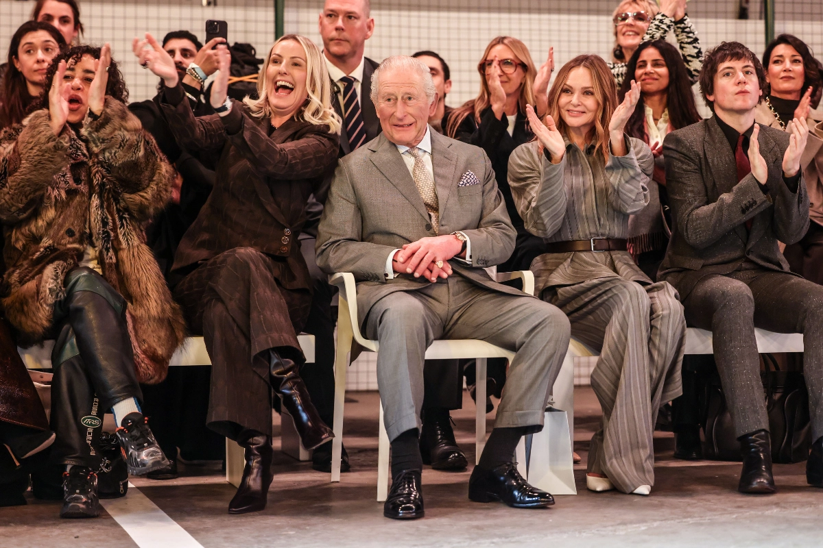 Londra Moda Haftası 2026 açılışında ön sırada oturan İngiltere Kralı Charles, gri takım elbisesiyle defileyi izlerken yanında stil sahibi konuklar alkış tutuyor; etkinliğin prestijli ve tarihi anını yansıtan front row görünümü.
