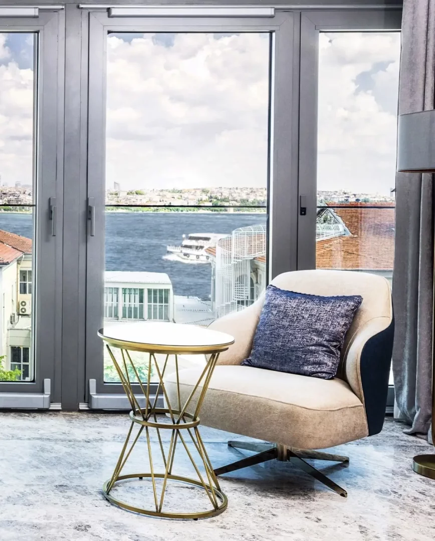 İstanbul'daki Maestro Hotel'in penceresinden Boğaz'ın ve karşı yakanın büyüleyici şehir manzarasına bakan, lacivert yastıklı krem rengi konforlu bir koltuk ile modern tasarımlı altın rengi sehpanın bulunduğu, şık ve ferah bir iç mekanın genel görünümü.