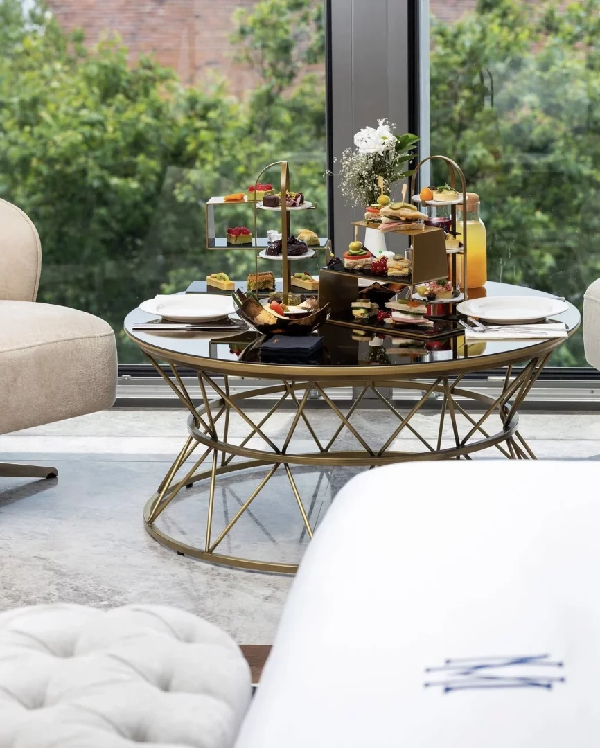 Maestro Hotel İstanbul'daki modern ve şık bir otel salonunda, geniş cam pencerelerden görünen yemyeşil ağaçların huzurlu manzarası eşliğinde, gold detaylı sehpa üzerinde üç katlı standlarda özenle sergilenen çeşit çeşit tatlı ve tuzlu ikramlarla dolu, davetkar bir ikindi çayı masası.
