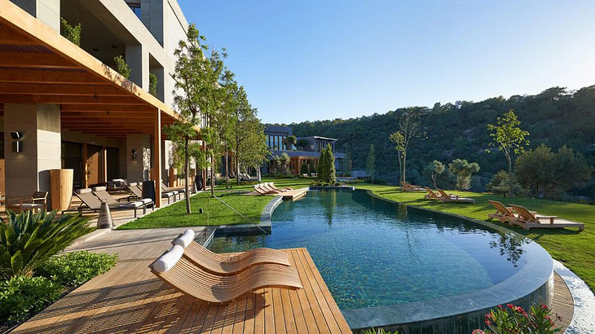 Mandarin Oriental Bodrum'da modern bir binanın önünde, ahşap şezlonglu bir güneşlenme terası ve yeşil çimlerle çevrili, doğal formuyla dikkat çeken geniş bir yüzme havuzu ve yemyeşil tepelerin yükseldiği bir manzara.