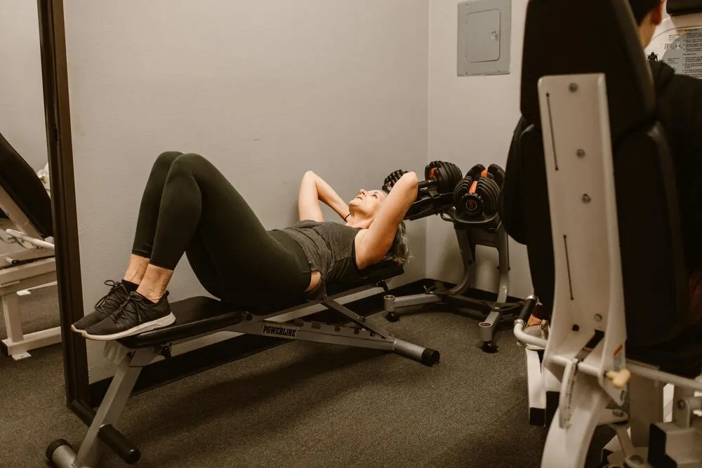 Spor salonunda bench üzerinde mekik çeken kadın; menopoz sonrası güçlenme ve core egzersizini temsil eden sahne.