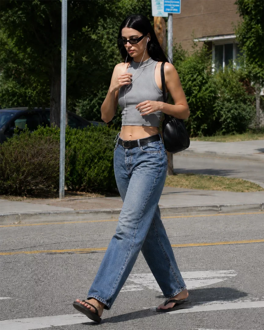Güneşli bir günde caddede yürüyen uzun siyah saçlı bir kadın, gri crop top'u ve beli açıkta bırakan rahat kesim düz paça jean'iyle, güncel jean trendleri arasında öne çıkan spor ve şık bir denim kombin örneği sergiliyor.