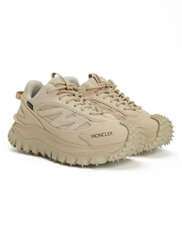 Bir çift Moncler bej renkli kadın sneaker ayakkabı, kalın ve tırtıklı tabanları ile ön kısmındaki belirgin dişli tasarımı sayesinde dikkat çekerken, süet ve sentetik deri panelleri modern ve dayanıklı bir görünüm sağlıyor.