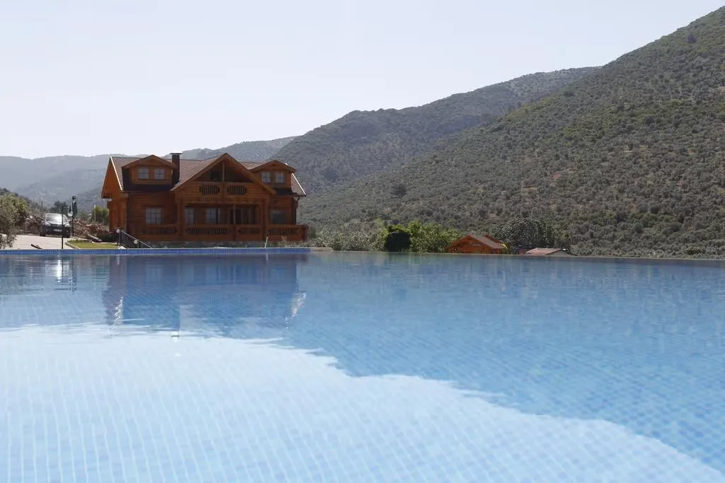Şirince'deki Natureland Efes Pansiyonu'nun sonsuzluk havuzunun berrak mavi mozaik karoları, misafirleri ferahlatıcı bir deneyime davet ederken, ardında yükselen yemyeşil ağaçlarla kaplı dağların ve ahşap iki katlı pansiyon binasının huzurlu yansımalarını sergiliyor.