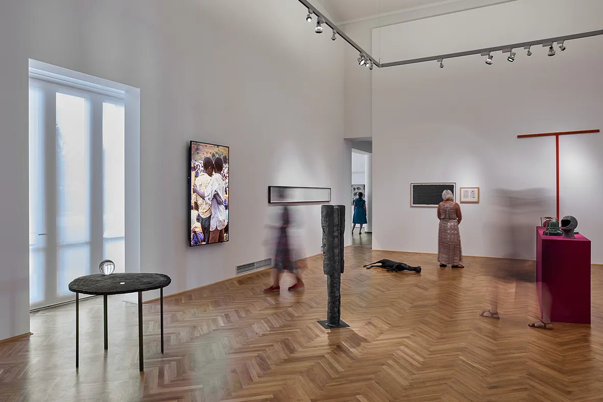 Aydınlık bir sanat galerisinde balıksırtı ahşap zeminde, duvarda çocuklar temalı bir fotoğraf sergisi, yerde soyut heykeller, pembe bir kaide üzerinde nesneler ve eserleri hareketli bir şekilde gezen ziyaretçiler görülüyor.