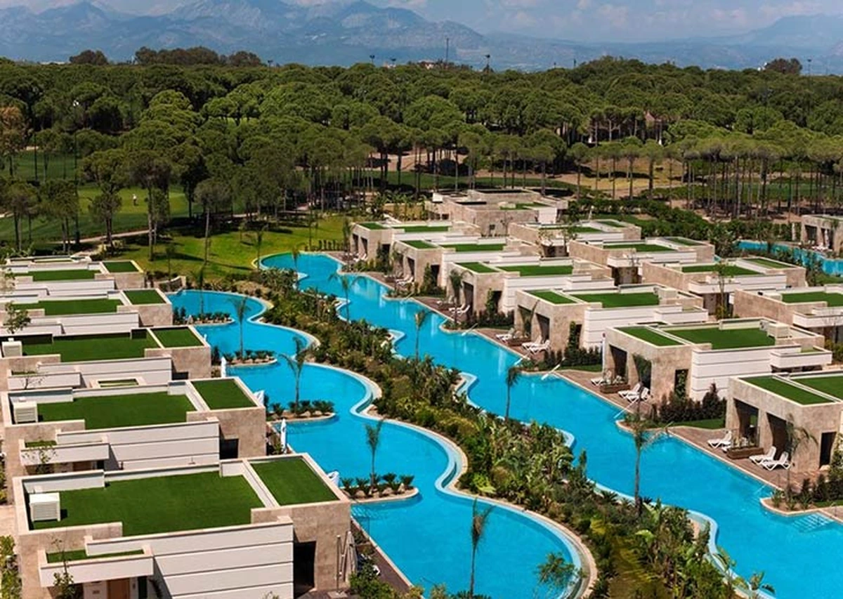 Regnum Carya Golf & Spa Resort'un geniş bir alanını gösteren kuşbakışı çekimde, yemyeşil çam ormanlarıyla çevrili golf sahasının yanı başında, birbirine bağlı turkuaz havuzlar boyunca sıralanmış, modern mimariye sahip ve çatılarında çim peyzajı bulunan çok sayıda villa, uzaktaki dağların fonunda lüks bir dinlenme tesisini gözler önüne seriyor.