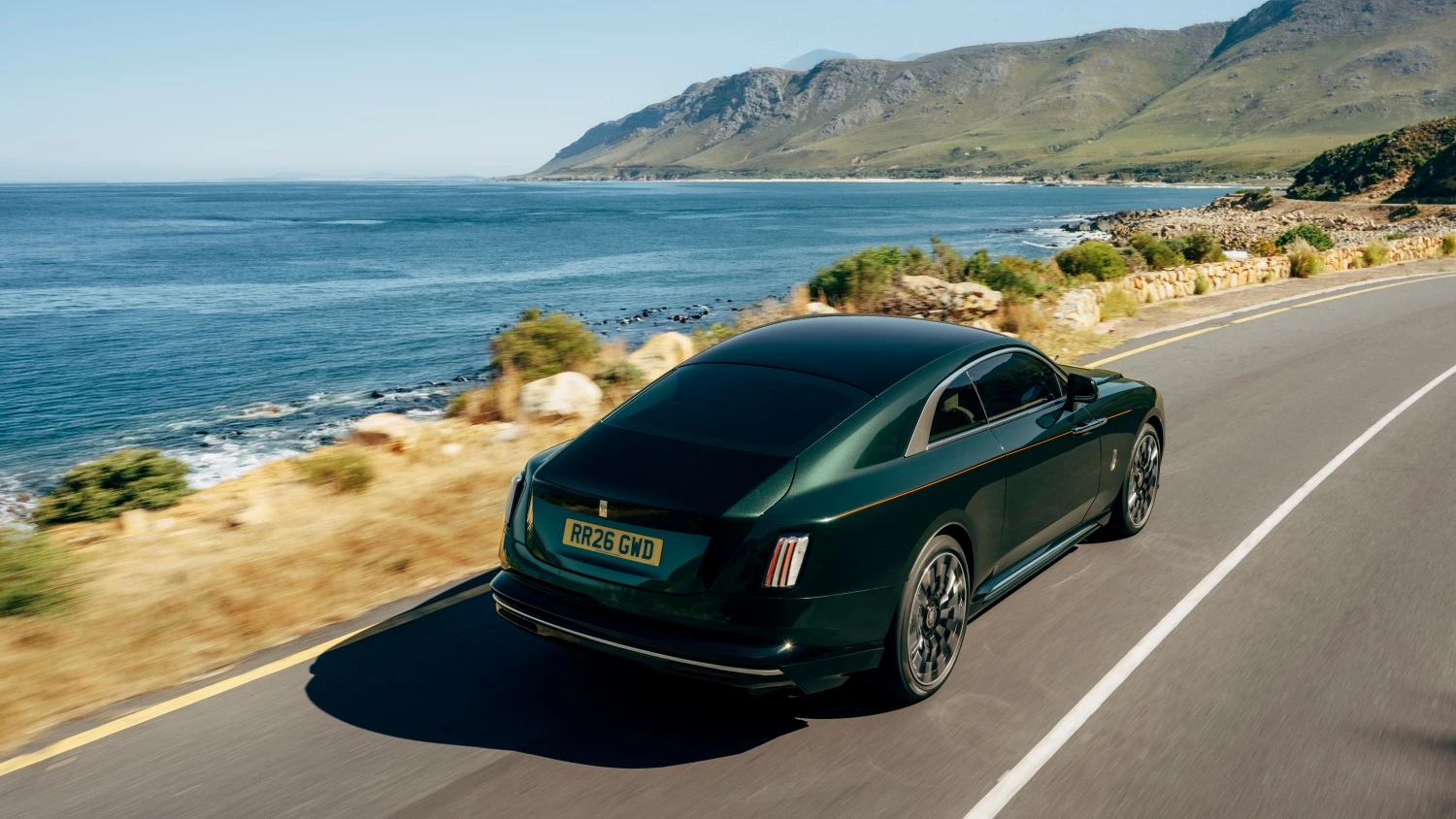 Mavi denizin ve yeşil dağların eşlik ettiği, güneşli bir sahil yolunda hızla ilerleyen koyu yeşil Rolls-Royce Spectre, lüks elektrikli otomobilin çağdaş ve makyajlı arka tasarımını sergiliyor.