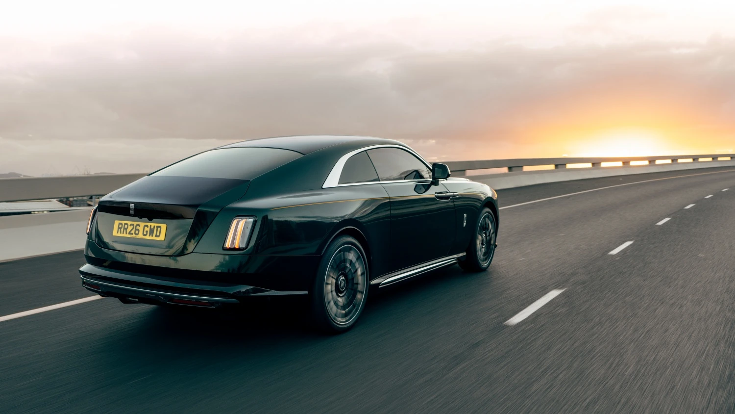 Zeytin yeşili rengiyle göz kamaştıran, makyajlı Rolls-Royce Spectre Series 2'nin yolda ilerlerken arka üç çeyrek görünümünü sergileyen bu fotoğrafta, aracın akıcı hatları ve yenilenen arka tasarım detayları gün batımının turuncu ışıkları altında modern bir şıklık sunuyor.