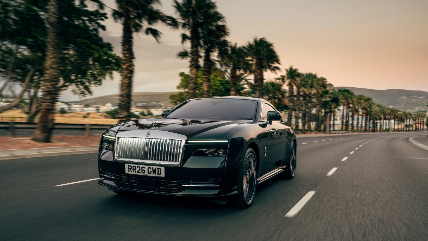 Makyajlı Rolls-Royce Spectre lüks elektrikli otomobil, palmiye ağaçları ve dağlarla çevrili, asfalt bir yolda gün batımının yumuşak ışıkları altında ilerlerken şık ve modern tasarımıyla dikkat çekiyor.