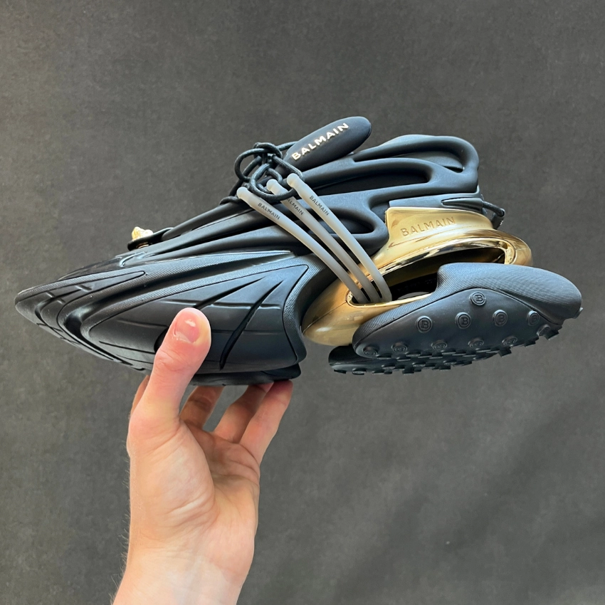 Siyah gövdeye ve altın detaylı tabana sahip Balmain futuristik sneaker, elde tutularak çekilmiş stüdyo çekimi.