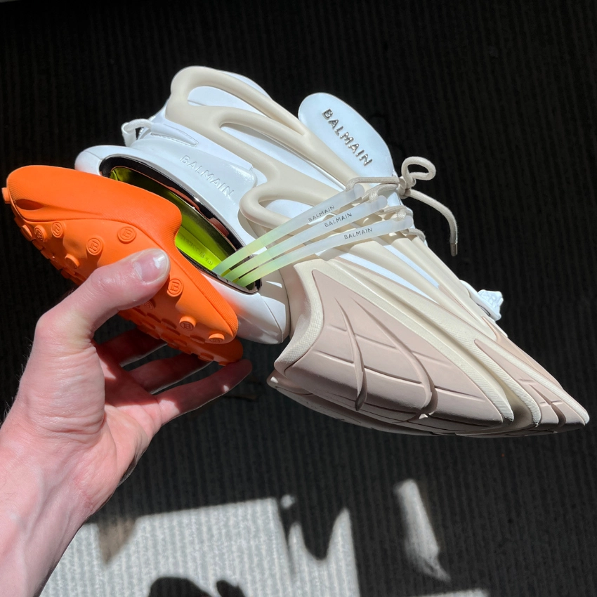 Turuncu taban detaylı, beyaz Balmain futuristik sneaker modeli, elde tutularak çekilmiş yan açı ürün fotoğrafı.