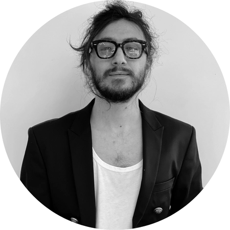 Siyah beyaz dairesel kadrajda, gözlüklü tasarımcı Safa Şahini blazer ceket ve tişörtle verdiği minimalist portre pozu.