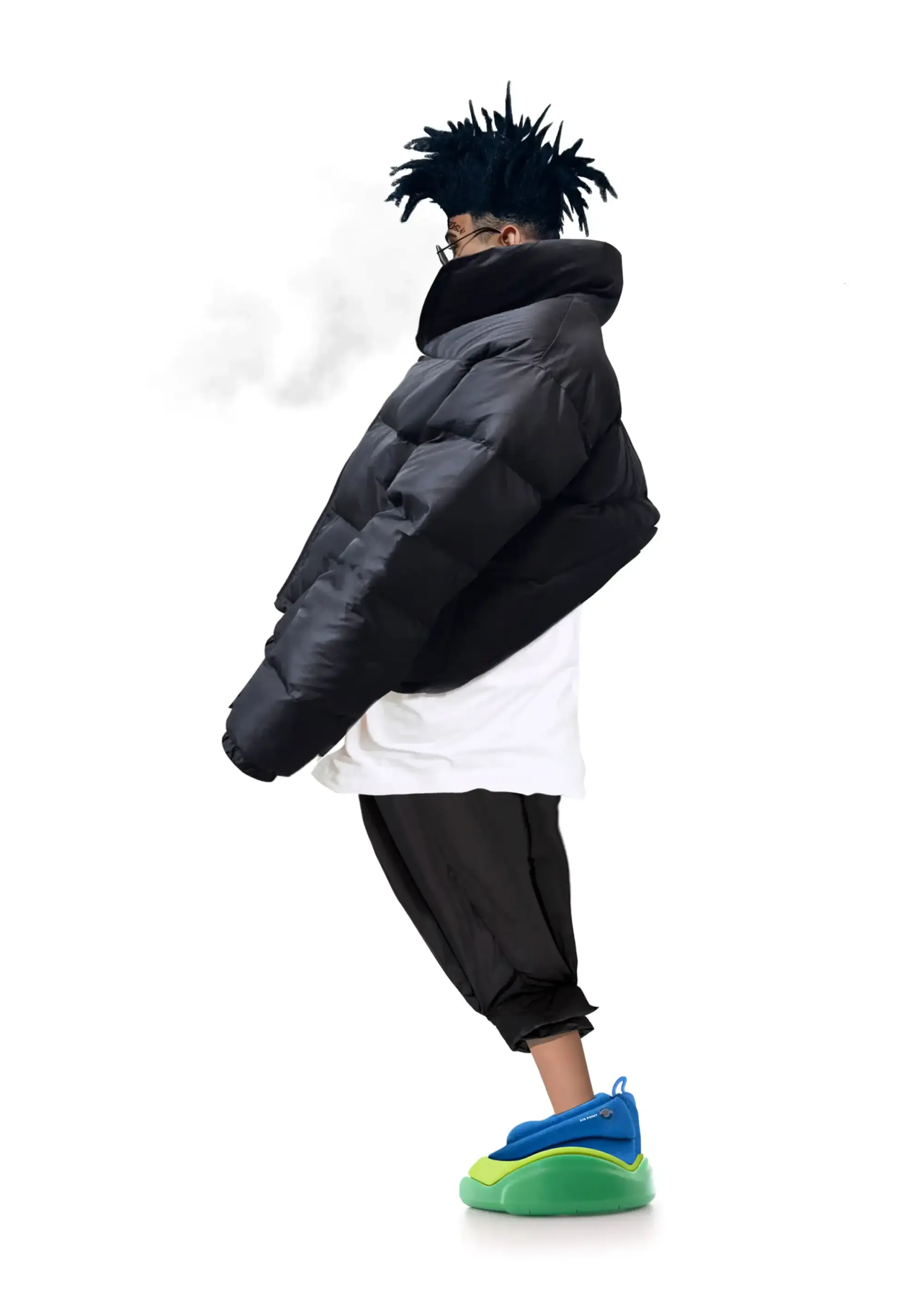 Siyah şişme mont ve oversize pantolonla poz veren model, mavi ve yeşil tabanlı futuristik sneaker tasarımıyla beyaz fonda yandan görünüm.